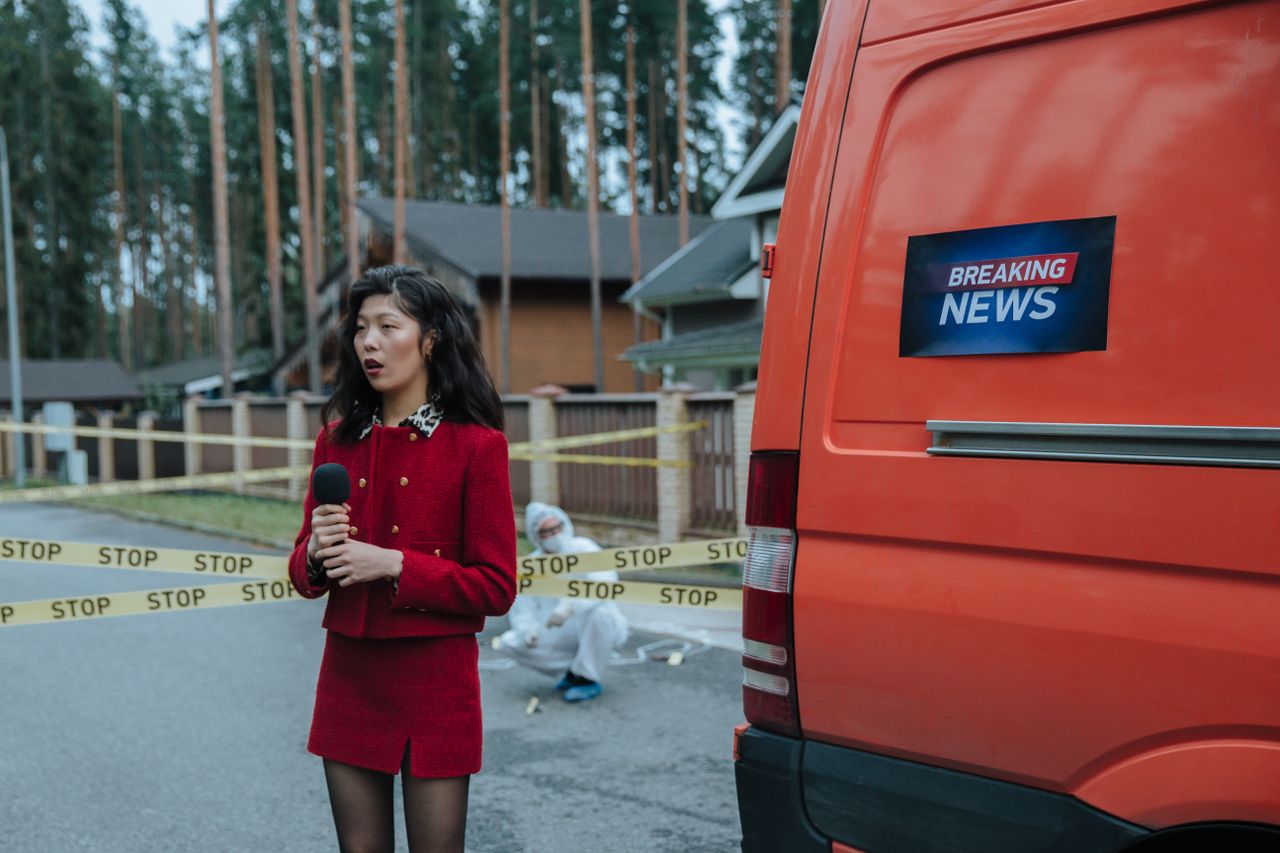 Female reporter in red outfit at an outdoor crime scene with warning tape and a news van.